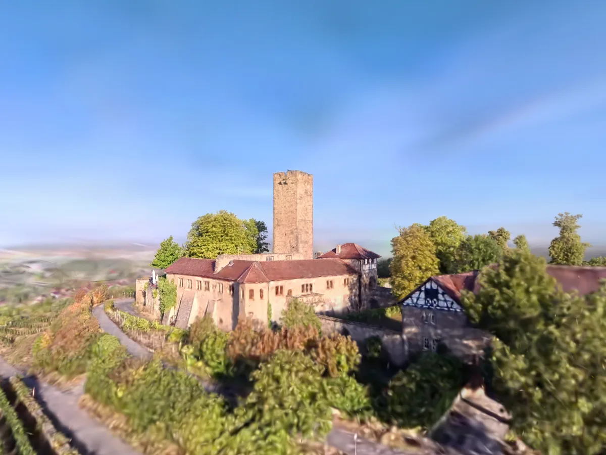 Schräge Luftaufnahme etwa auf Dachhöhe unter weitem, dunstigem Himmel — in der Bildmitte der quadratische Bergfried aus hellem Sandstein, dachlos und oben ausgebrochen; davor die zweigeschossigen Bauten der Kernburg mit roten Ziegeldächern und Reihen rechteckiger Fenster, an ihrem linken Ende ein geschlossener weißer Sonnenschirm auf der Terrasse. Rechts ein Fachwerkbau über steinernem Erdgeschoss, dahinter läuft eine Mauer weiter. Bäume säumen die Anlage auf beiden Seiten, davor fallen terrassierte Weinberge ab, durch die sich ein Fahrweg windet; links unten die roten Dächer von Sulzfeld und dahinter die dunstige Ebene.